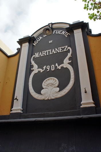An architectural element with ornate lettering and decorative designs features the words 'AGUA DE FUENTE DE MARTIANEZ 1901.' The structure is flanked by columns, with decorative moldings and a shell motif at the center. There is a small faucet on one side.
