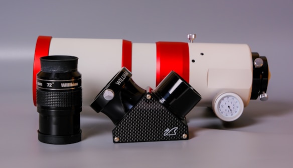 A telescope and its components are prominently displayed. The main telescope tube is white with red accents and lies horizontally. A black eyepiece labeled 'William 72°' is positioned to the side along with another black attachment. A gauge is affixed to the side of the telescope.