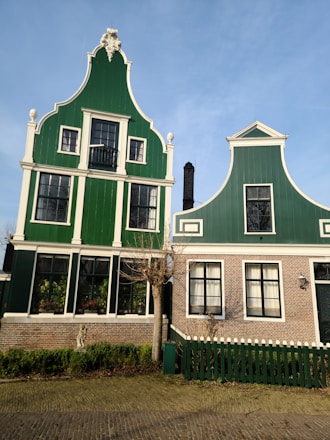 Two traditional Dutch-style houses with ornate architectural details and green wooden facades. The left house features a decorative gable with statuesque detailing at the top, and multiple windows with white trim. A bare tree is positioned in front of the houses, alongside neatly trimmed hedges.