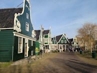 A row of green, traditional Dutch houses with ornate gables line a cobblestone street. These buildings have white decorative trim and steep, dark-tiled roofs. A few people are walking and standing in the area, some near the houses and a few on the path. There is a leafless tree on the right side, and the sky is clear with a gentle blue hue.
