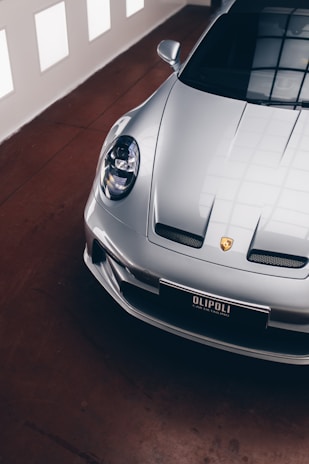 A sleek, silver luxury sports car is parked indoors on a polished brown floor. The image shows a partial view focused on the front and side of the car, highlighting its aerodynamic design and distinct headlight. The car's emblem and a license plate with the text 'OLIPOLI CAR DETAILING' are visible. The lighting creates soft reflections on the car's surface, enhancing its glossy finish. Large windows in the background allow natural light to enter, creating a mix of shadows and reflections on the floor.
