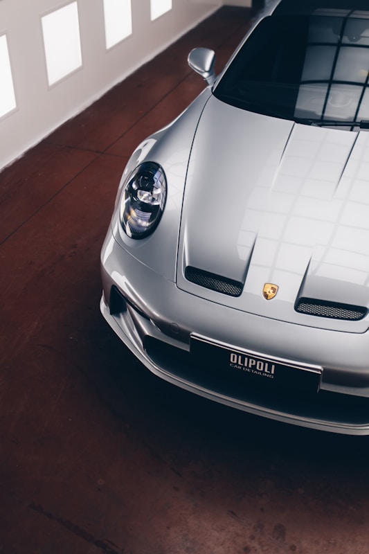 A sleek, silver luxury sports car is parked indoors on a polished brown floor. The image shows a partial view focused on the front and side of the car, highlighting its aerodynamic design and distinct headlight. The car's emblem and a license plate with the text 'OLIPOLI CAR DETAILING' are visible. The lighting creates soft reflections on the car's surface, enhancing its glossy finish. Large windows in the background allow natural light to enter, creating a mix of shadows and reflections on the floor.