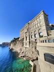 A grand, historic building with ornate architecture is perched on a rocky cliff overlooking the clear, turquoise sea. The sky is a vibrant blue, and the sunlight casts shadows on the building's stone facade. In the distance, other buildings dot the coastline.