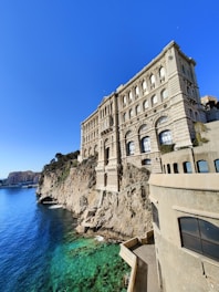 A grand, historic building with ornate architecture is perched on a rocky cliff overlooking the clear, turquoise sea. The sky is a vibrant blue, and the sunlight casts shadows on the building's stone facade. In the distance, other buildings dot the coastline.