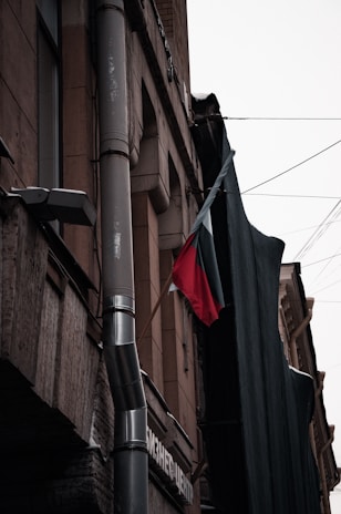 A multi-story building facade is shown with pipes and a protruding window. A part of a banner or large black cloth is hanging alongside the structure. Below, a red and black flag is attached to the building. The structure appears to be made of stone or concrete with metallic elements and the atmosphere is overcast, resulting in muted lighting.