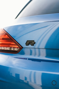 A close-up view of a blue car's rear section, featuring a stylish R emblem prominently displayed. The tail light is partially visible with a sleek design and red coloration. The background shows a soft reflection on the car's surface, suggesting a bright sunny day.