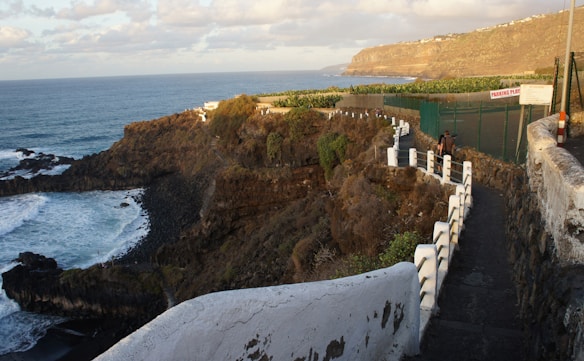 A scenic coastal pathway with a view of the ocean meeting rocky cliffs. The path is bordered by a white railing, and vegetation covers the cliffs. The sky is partly cloudy, and there is a fenced area with a sign visible near the path.