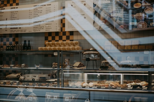 A bakery display with various pastries and desserts arranged on shelves behind a glass counter. There are several different baked goods, such as croissants and cakes, some under glass domes. The background features a menu board with items listed in both English and another language. There are decorative bottles and other items on the upper shelves.