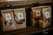 Paper bags with transparent windows are arranged on a wooden shelf, each labeled with product names such as Madeleine and Chestnuts, accompanied by price tags in both English and Chinese.