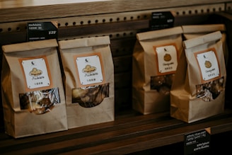 Paper bags with transparent windows are arranged on a wooden shelf, each labeled with product names such as Madeleine and Chestnuts, accompanied by price tags in both English and Chinese.