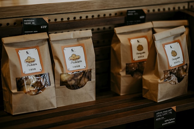 Paper bags with transparent windows are arranged on a wooden shelf, each labeled with product names such as Madeleine and Chestnuts, accompanied by price tags in both English and Chinese.