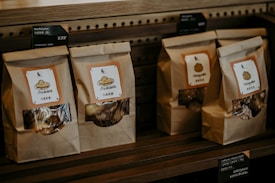 Paper bags with transparent windows are arranged on a wooden shelf, each labeled with product names such as Madeleine and Chestnuts, accompanied by price tags in both English and Chinese.