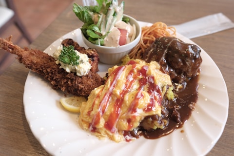 A plate of assorted food items, including a piece of fried shrimp with tartar sauce and a parsley garnish, an omelet drizzled with ketchup, a hamburger steak covered in brown gravy, and a small bowl of salad with mixed greens and dressing. The plate also includes a small serving of spaghetti.