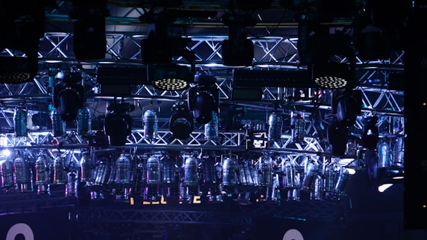 A complex network of metal trusses supporting numerous stage lights and equipment, with a prominent use of blue and purple lighting creating a futuristic atmosphere.