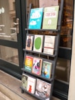 A black wooden shelf stands outside a shop, displaying a variety of books with colorful covers. The books are arranged in four rows, each holding three to four books, featuring different titles and designs. The shop door, visible to the left, has some writing on it including the contact details for the shop.