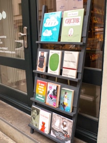 A black wooden shelf stands outside a shop, displaying a variety of books with colorful covers. The books are arranged in four rows, each holding three to four books, featuring different titles and designs. The shop door, visible to the left, has some writing on it including the contact details for the shop.