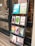 A black wooden shelf stands outside a shop, displaying a variety of books with colorful covers. The books are arranged in four rows, each holding three to four books, featuring different titles and designs. The shop door, visible to the left, has some writing on it including the contact details for the shop.