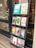 A black wooden shelf stands outside a shop, displaying a variety of books with colorful covers. The books are arranged in four rows, each holding three to four books, featuring different titles and designs. The shop door, visible to the left, has some writing on it including the contact details for the shop.
