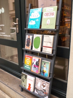 A black wooden shelf stands outside a shop, displaying a variety of books with colorful covers. The books are arranged in four rows, each holding three to four books, featuring different titles and designs. The shop door, visible to the left, has some writing on it including the contact details for the shop.