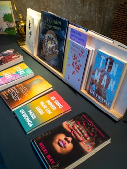 A display of various books arranged on a table, with some books standing upright in a wooden holder and others lying flat. The covers feature a mix of bright colors, illustrations, and text. A brass lamp and a woven basket add to the display decor.