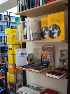 A variety of books are arranged on shelves in a bookstore. Some books are stacked vertically while others are displayed horizontally. Prominent are yellow books with a photo of a woman on the cover. Other books feature different designs and colors. The bookshelves are well-organized, showcasing a diverse collection.