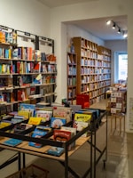 A cozy bookstore interior with shelves filled with a variety of books. The image features two main sections: on the left, several black metal shelves filled with colorful children's books, visible with titles on the front. To the right, long rows of wooden bookshelves extend into the background, filled with books of different sizes and colors. A red armchair is tucked into a reading nook near the back, and there's soft overhead lighting providing a warm ambiance.