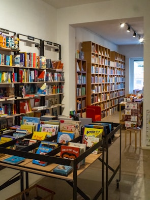 A cozy bookstore interior with shelves filled with a variety of books. The image features two main sections: on the left, several black metal shelves filled with colorful children's books, visible with titles on the front. To the right, long rows of wooden bookshelves extend into the background, filled with books of different sizes and colors. A red armchair is tucked into a reading nook near the back, and there's soft overhead lighting providing a warm ambiance.