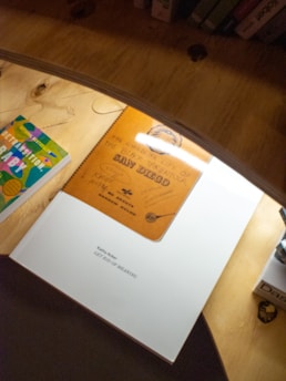 A close-up view of books on a wooden surface, including a colorful book on the left and a large white book with the title 'GET RID OF MEANING' at the center. A brown book labeled 'San Diego' with handwritten text is also visible, partially overlapping the white book.