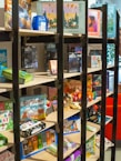 A bookshelf contains a variety of board games, puzzles, and card games. The shelves display colorful and diverse game boxes, with some dedicated to adventure themes. The scene is lively and inviting, with a mix of cartoonish and realistic illustrations on the packaging.