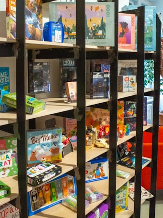 A bookshelf contains a variety of board games, puzzles, and card games. The shelves display colorful and diverse game boxes, with some dedicated to adventure themes. The scene is lively and inviting, with a mix of cartoonish and realistic illustrations on the packaging.
