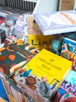A collection of colorful books is stacked on a table. The covers feature vibrant designs and images related to cuisine, with prominent titles in various languages. The background includes more books and a section of a shelf.