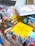 A collection of colorful books is stacked on a table. The covers feature vibrant designs and images related to cuisine, with prominent titles in various languages. The background includes more books and a section of a shelf.
