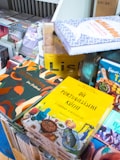 A collection of colorful books is stacked on a table. The covers feature vibrant designs and images related to cuisine, with prominent titles in various languages. The background includes more books and a section of a shelf.