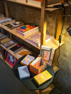 A wooden shelving unit filled with various books and colorful boxes. On the top shelf, there is a desk lamp attached to the side. The middle shelf hosts several books stacked both vertically and horizontally. The bottom shelf features a few more books and magazines. The table in front holds a few open and closed boxes with vibrant designs.