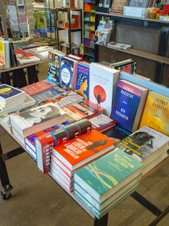 A display table filled with a variety of books in a bookstore. The books are stacked and arranged to showcase their covers. Shelves and other merchandise are visible in the background, along with a colorful assortment of items.