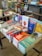 A display table filled with a variety of books in a bookstore. The books are stacked and arranged to showcase their covers. Shelves and other merchandise are visible in the background, along with a colorful assortment of items.