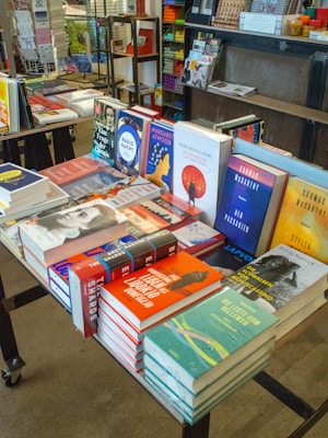 A display table filled with a variety of books in a bookstore. The books are stacked and arranged to showcase their covers. Shelves and other merchandise are visible in the background, along with a colorful assortment of items.