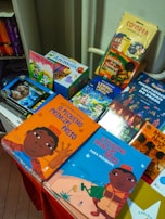 A colorful display of children's books is arranged on a table, featuring vibrant covers with illustrations. The books include titles in both Portuguese and English, with some books showcasing diverse cultural themes and characters.