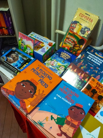 A colorful display of children's books is arranged on a table, featuring vibrant covers with illustrations. The books include titles in both Portuguese and English, with some books showcasing diverse cultural themes and characters.