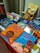 A colorful display of children's books is arranged on a table, featuring vibrant covers with illustrations. The books include titles in both Portuguese and English, with some books showcasing diverse cultural themes and characters.