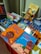 A colorful display of children's books is arranged on a table, featuring vibrant covers with illustrations. The books include titles in both Portuguese and English, with some books showcasing diverse cultural themes and characters.