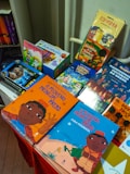 A colorful display of children's books is arranged on a table, featuring vibrant covers with illustrations. The books include titles in both Portuguese and English, with some books showcasing diverse cultural themes and characters.