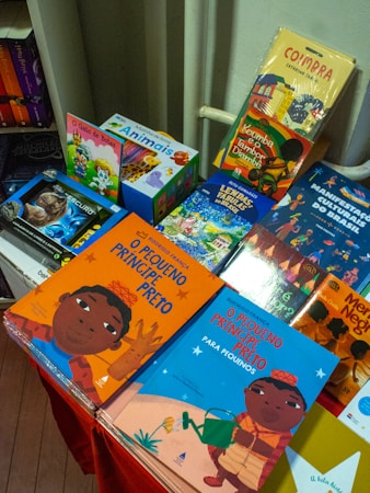 A colorful display of children's books is arranged on a table, featuring vibrant covers with illustrations. The books include titles in both Portuguese and English, with some books showcasing diverse cultural themes and characters.