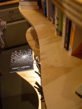 A cozy indoor setting with a wooden bookshelf partially filled with books. A single book with a dark cover titled 'Treieben Lassan' is resting on the floor. The room appears warmly lit, and there are some drawers with handles visible.