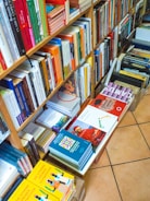 The image displays a well-organized bookshelf filled with a variety of colorful books. Books are stacked vertically on shelves, and there are also piles of books placed horizontally on the floor, organized in boxes. The titles and spines of the books exhibit a range of colors, including reds, oranges, yellows, blues, and greens.