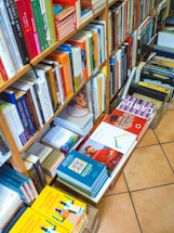 The image displays a well-organized bookshelf filled with a variety of colorful books. Books are stacked vertically on shelves, and there are also piles of books placed horizontally on the floor, organized in boxes. The titles and spines of the books exhibit a range of colors, including reds, oranges, yellows, blues, and greens.