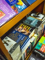 A bookshelf filled with various books, including titles in different languages. Some visible books include 'Mulheres que Correm com os Lobos' and 'Clarice'. The books are stacked in an orderly manner, with some lying flat and others standing upright. The shelves are wooden, adding a warm tone to the setting.