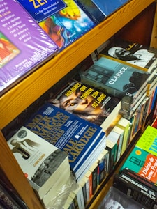 A bookshelf filled with various books, including titles in different languages. Some visible books include 'Mulheres que Correm com os Lobos' and 'Clarice'. The books are stacked in an orderly manner, with some lying flat and others standing upright. The shelves are wooden, adding a warm tone to the setting.