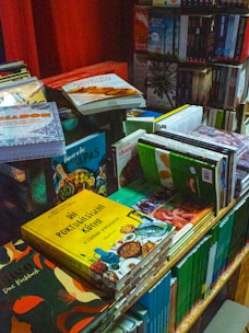 A cluttered and vibrant bookstand filled with a variety of cookbooks and novels. The books are stacked in different orientations, some are upright while others are laid flat. The covers display bright colors and images, particularly focusing on cuisines with titles in different languages, including Portuguese and German.