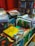 A cluttered and vibrant bookstand filled with a variety of cookbooks and novels. The books are stacked in different orientations, some are upright while others are laid flat. The covers display bright colors and images, particularly focusing on cuisines with titles in different languages, including Portuguese and German.